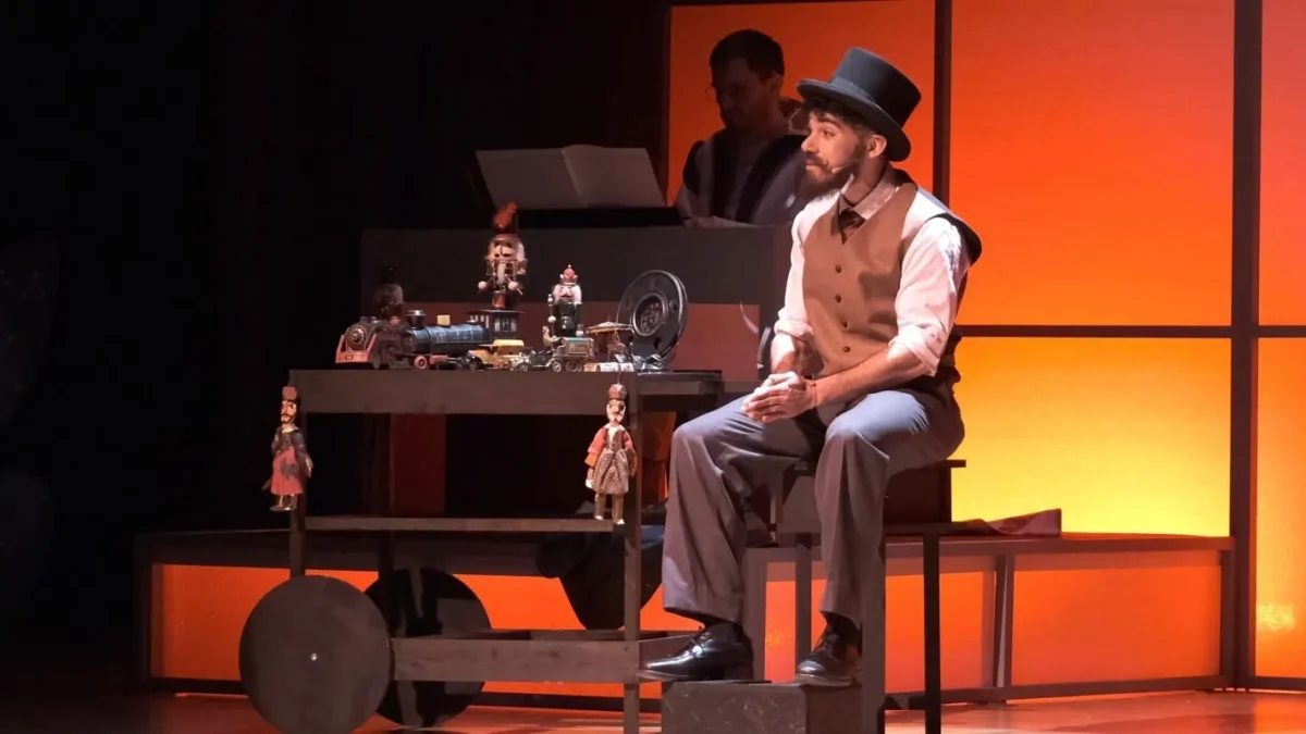 Actor en medio del escenario, con ropa antigüa, sombrero de bombin, sobre un taburete de madera y con dos marionetas.