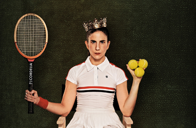 Una chica posada sentada en una silla con corona de reina, vestida de tenista, en una mano sujeta la raqueta y en otra 3 pelotas de tenis
