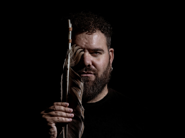 Abraham Cupeiro en primer plano con fondo negro y cubriendose media cara con un instrumento musical que tiene una pluma.